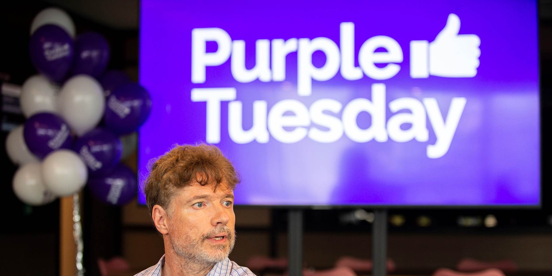 A picture of Purple Tuesday's CEO Mike, a man of small stature standing in front of a Purple Tuesday sign