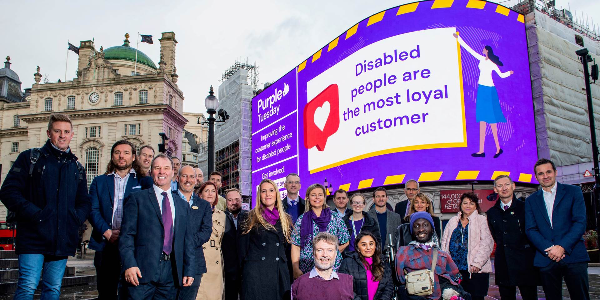 A group photo taken in front of the Piccadilly Lights featuring the sponsors of Purple Tuesday 2022, Purple Tuesday CEO Mike Adams, and Purple Tuesday ambassadors. The individuals are posing together, with the Piccadilly Lights prominently displaying the 
